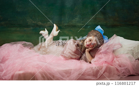Graceful Rest. Elegant young woman, royal person lying on bed with pink veiled and smiling against vintage green background. Historical charm 116801217