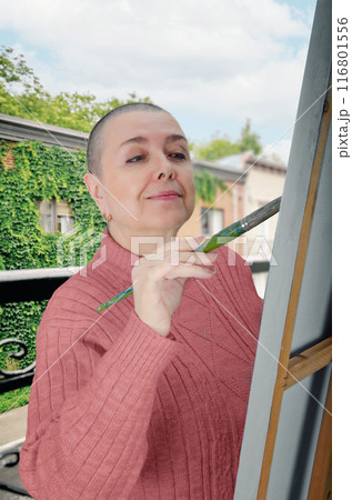 Shaved-haired woman undergoing chemotherapy paints on an easel, using art therapy to restore her color perception and fine motor skills. 116801556