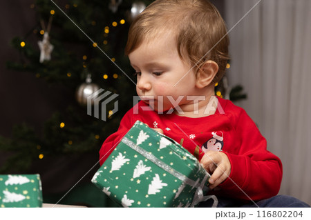 A one-year-old kid in a Santa hat opens gifts near the Christmas tree 116802204