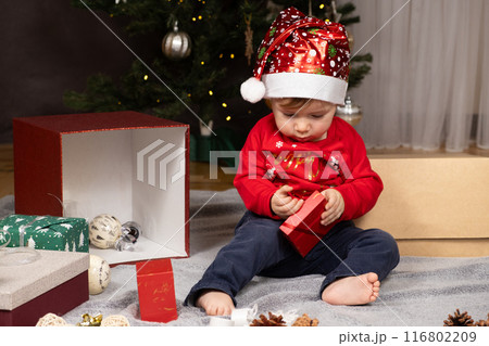 A one-year-old kid in a Santa hat opens gifts near the Christmas tree 116802209