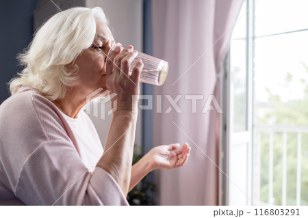 Senior woman takes pill with glass of water in hand.  116803291