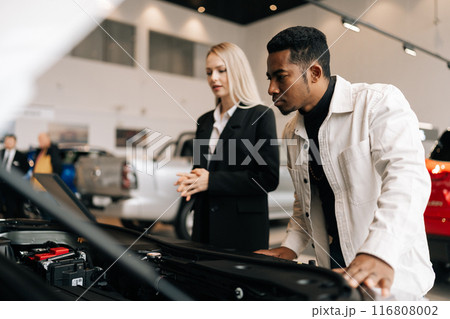 Female sales manager and African male client checking motor under bonnet talking gesturing in showroom. Focused young black businessman choosing new automobile in dealership office. 116808002