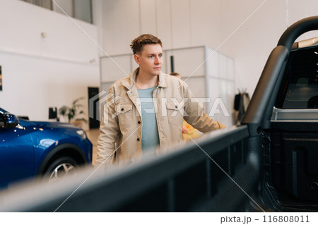 Young man buyer looking around big open trunk of car. Male customer choosing pickup truck in car dealership. Guy visitor walking in modern auto showroom and choosing new vehicle to buy or rent. 116808011