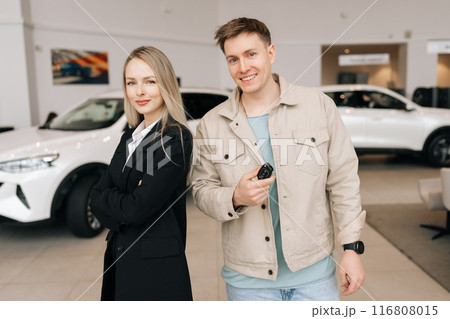 Portrait of satisfied young man buyer holding in hand keys of new car standing posing with friendly blond saleswoman in suit in bright showroom, smiling looking at camera. Concept of buying vehicle. Portrait of satisfied young man buyer holding in hand keys of new car standing posing with friendly blond saleswoman in suit in bright showroom, smiling looking at camera. Concept of buying vehicle. 116808015