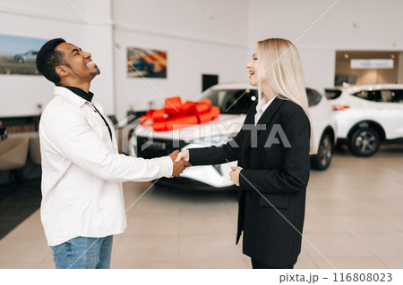 Happy excited African-American buyer getting car key and greetings from female auto dealer while buying new auto in dealership. Smiling black car owner during handing over keys at auto showroom. Happy excited African-American buyer getting car key and greetings from female auto dealer while buying new auto in dealership. Smiling black car owner during handing over keys at auto showroom. 116808023