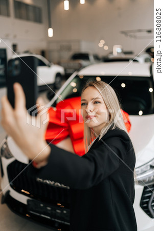Vertical shot of happy female customer in black suit doing selfie shot pov on mobile phone choose auto want to buy new automobile in car showroom. Concept of choosing and buying new auto at showroom. 116808025