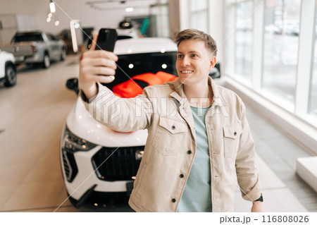 Cheerful male customer in casual clothes taking selfie on mobile phone near new car at auto dealership. Joyful handsome buyer photographing at showroom, buying automobile. Concept of buying new auto. Cheerful male customer in casual clothes taking selfie on mobile phone near new car at auto dealership. Joyful handsome buyer photographing at showroom, buying automobile. Concept of buying new auto. 116808026