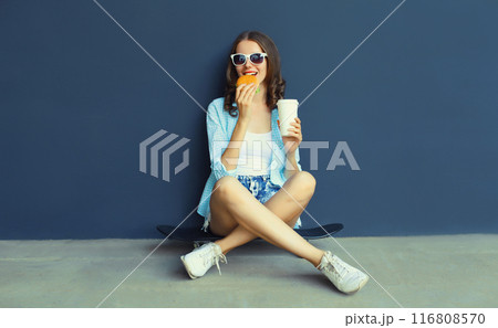 Happy modern young woman eating burger fast food, stylish teenage girl sitting on skateboard in city 116808570
