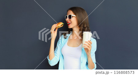 Happy cheerful young woman eating burger fast food holding cup of coffee, teenage girl having fun Happy cheerful young woman eating burger fast food holding cup of coffee, teenage girl having fun 116808573