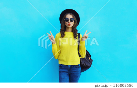 Portrait of stylish young woman in black hat, yellow sweater, modern cool girl posing on blue 116808586