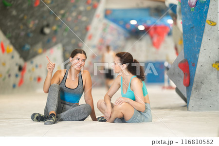 Two athletic women sit on floor and discuss options for climbing a climbing wall without special equipment 116810582