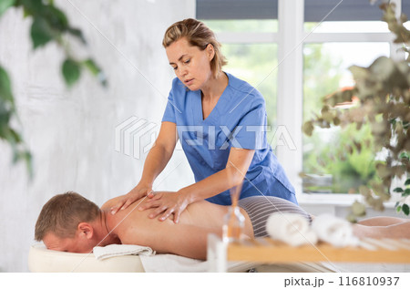 Female masseur performs back stroking and patting during massage session for male client Female masseur performs back stroking and patting during massage session for male client 116810937
