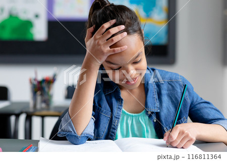 Happy biracial schoolgirl sitting at desk in class, resting head on hand and writing, copy space Happy biracial schoolgirl sitting at desk in class, resting head on hand and writing, copy space 116811364