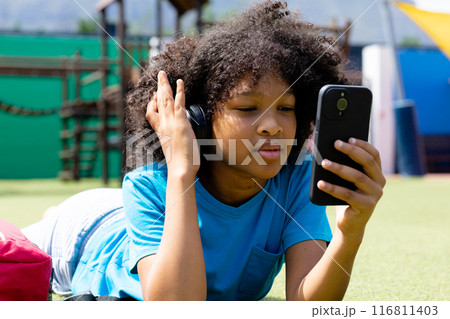 Happy biracial schoolgirl in headphones using smartphone, lying in schoolyard, copy space Happy biracial schoolgirl in headphones using smartphone, lying in schoolyard, copy space 116811403