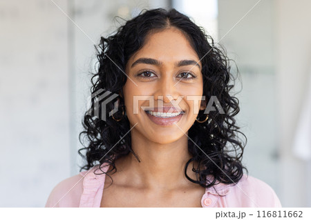 Smiling woman with curly hair wearing pink shirt, looking at camera indoors Smiling woman with curly hair wearing pink shirt, looking at camera indoors 116811662