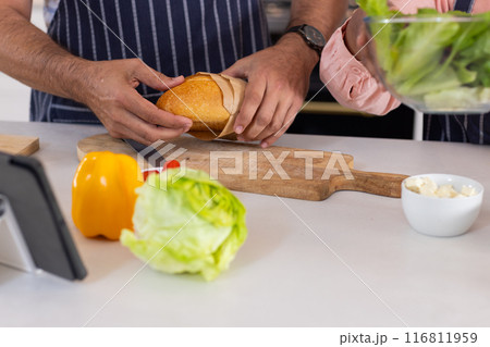Cutting bread on wooden board, senior couple preparing fresh salad in kitchen 116811959