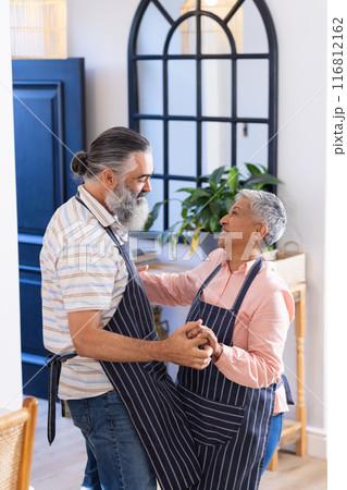 Senior couple dancing together in kitchen, wearing aprons and smiling joyfully Senior couple dancing together in kitchen, wearing aprons and smiling joyfully 116812162