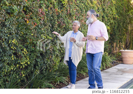 Senior couple walking and discussing garden plants while holding coffee cups, copy space Senior couple walking and discussing garden plants while holding coffee cups, copy space 116812255
