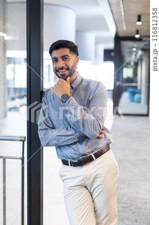 Leaning against glass wall, smiling businessman in office wearing smart casual attire Leaning against glass wall, smiling businessman in office wearing smart casual attire 116812358