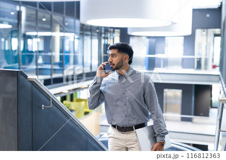 Businessman walking in office, talking on smartphone, wearing formal attire Businessman walking in office, talking on smartphone, wearing formal attire 116812363
