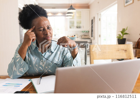 Busy senior african american woman with documents on table using laptop at home 116812511