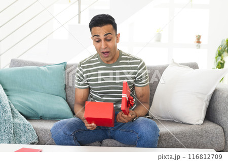 Middle Eastern man looks surprised during a video call as he opens a gift at home 116812709