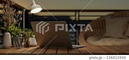 Back view image of a laptop computer on a wooden desk on a balcony at dusk, illuminated by a lamp. 116812930