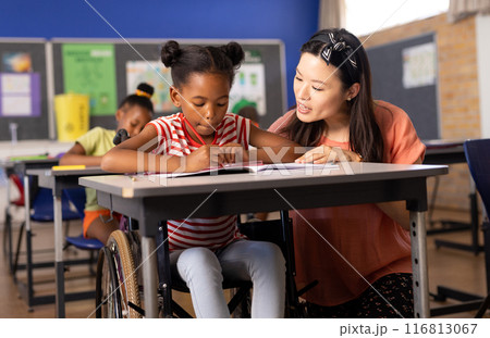 Asian female teacher assisting african american schoolgirl sitting on wheelchair to write notes 116813067