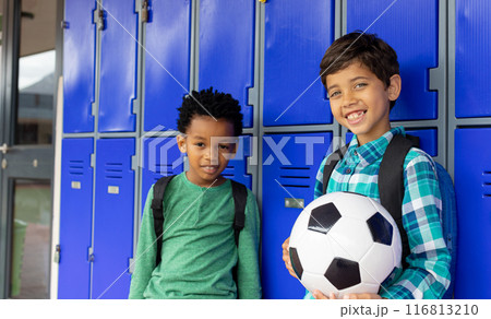 Biracial boy and African American boy stand smiling by school lockers, one holding a soccer ball 116813210