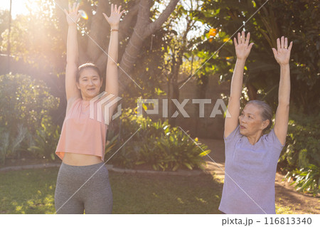 Exercising outdoors, asian grandmother and granddaughter stretching arms up in sunny garden Exercising outdoors, asian grandmother and granddaughter stretching arms up in sunny garden 116813340