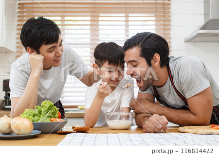 Happy cheerful gay couple with their adopted little son enjoy making and eating a sandwich with fresh milk for breakfast in the kitchen together. Happy LGBTQ+ family with adopted child concept. Happy cheerful gay couple with their adopted little son enjoy making and eating a sandwich with fresh milk for breakfast in the kitchen together. Happy LGBTQ+ family with adopted child concept. 116814422
