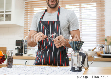 Happy handsome Asian man is using hand coffee grinder grinds a coffee beans. Man making a coffee at home in warmed atmosphere kitchen. 116814497