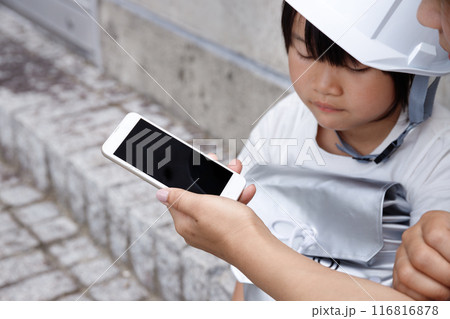 ヘルメットをかぶった小学生の女の子とママがスマホを操作。防災のイメージで安否確認をしている ヘルメットをかぶった小学生の女の子とママがスマホを操作。防災のイメージで安否確認をしている 116816878