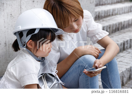 ヘルメットをかぶった小学生の女の子とママがスマホを操作。防災のイメージで安否確認をしている 116816881