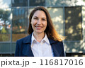 Portrait of a happy caucasian brunette woman in a blue suit stands against the background of a business building and smiles 116817016