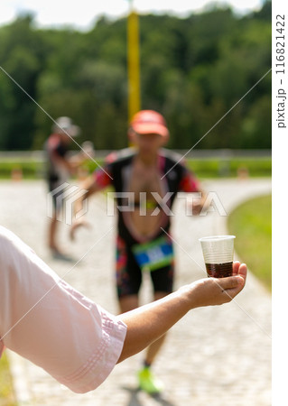 A volunteer in a white dress holds out a plastic cup of water to a triathlon runner. A volunteer in a white dress holds out a plastic cup of water to a triathlon runner. 116821422