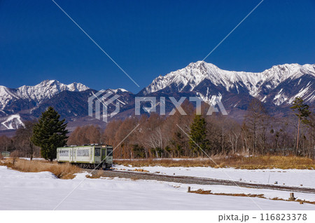 雪の八ヶ岳を背景に快晴の高原を行く小海線キハ110系 116823378