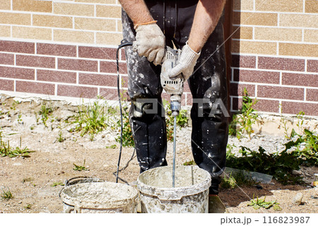 Construction worker stirs cement adhesive mortar to cover facade with flexible faux brick panels. Construction worker stirs cement adhesive mortar to cover facade with flexible faux brick panels. 116823987