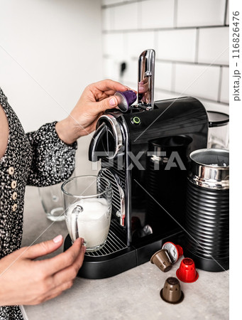 Girl with capsule coffee machine and milk in transparent cup preparing cappuccino at kitchen. Woman preparing italian caffeine beverage using professional espresso maker 116826476