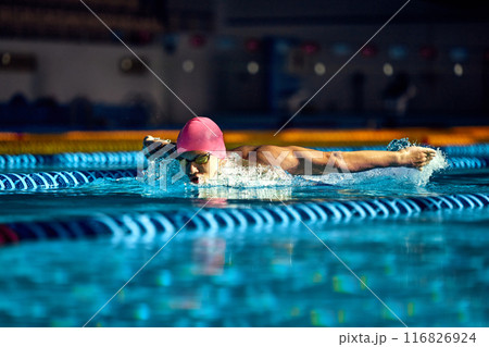 Athlete in pink cap and goggles executes butterfly stroke, his muscular arms cutting through water, creating intense and focused action shot in blue pool. Athlete in pink cap and goggles executes butterfly stroke, his muscular arms cutting through water, creating intense and focused action shot in blue pool. 116826924