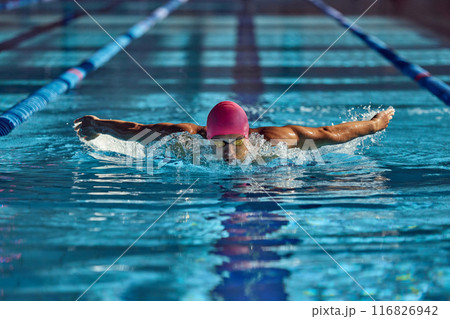 Man, swimmer in pink cap and goggles powers through water with butterfly stroke, his focused expression and strong arms creating dynamic splash in pool. Man, swimmer in pink cap and goggles powers through water with butterfly stroke, his focused expression and strong arms creating dynamic splash in pool. 116826942