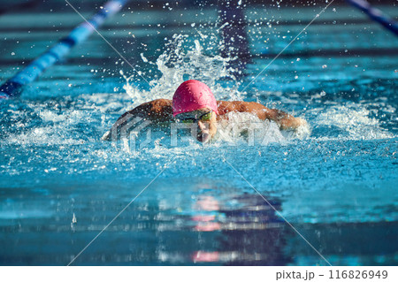 Man, swimmer in pink cap and goggles powers through water with butterfly stroke, his focused expression and strong arms creating dynamic splash in pool. 116826949