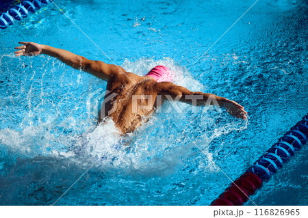 Overhead view of athlete in pink swim cap performing butterfly stroke, showcasing strength and technique as he propel through vibrant blue pool. 116826965