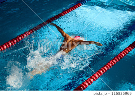 Swimmer in pink cap executes powerful butterfly stroke, his muscular form cutting through blue water, creating dynamic splash between red lane dividers. Swimmer in pink cap executes powerful butterfly stroke, his muscular form cutting through blue water, creating dynamic splash between red lane dividers. 116826969