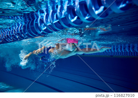 Competitive swimmer, with pink cap and blue trunks, seen underwater during dive, demonstrates his strength and form creating water turbulence as he moves forward. Competitive swimmer, with pink cap and blue trunks, seen underwater during dive, demonstrates his strength and form creating water turbulence as he moves forward. 116827010