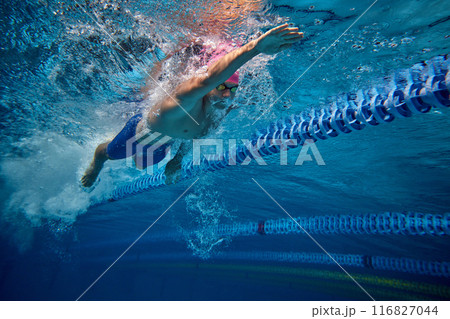 Male athlete swimming, highlighting his focus and form, as he pushes through water in pool, wearing blue swim trunks and goggles. 116827044