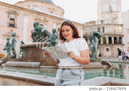 Attractive female tourist is exploring new city. Redhead happy woman holding a paper map near Fountain Rio Turia on Square of the Virgin Saint Mary, Valencia Cathedral. Traveling Europe 116827639