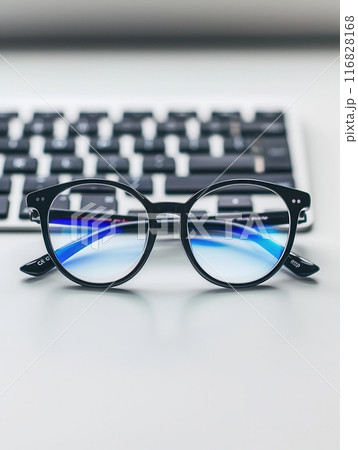 A pair of black eyeglasses sits on a white desk with a computer keyboard slightly out of focus in the background. 116828168