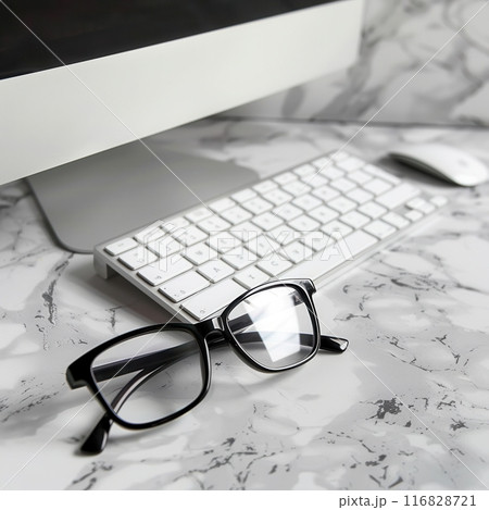 A pair of black eyeglasses rests on a white desk near a computer keyboard and monitor. The surface is textured like marble. A pair of black eyeglasses rests on a white desk near a computer keyboard and monitor. The surface is textured like marble. 116828721