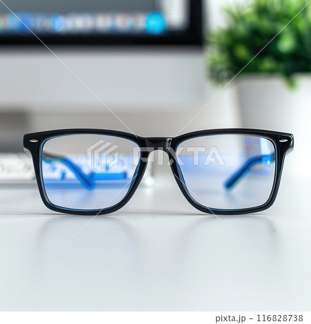A pair of black eyeglasses rest on a white desk with a computer screen and keyboard out of focus in the background. 116828738